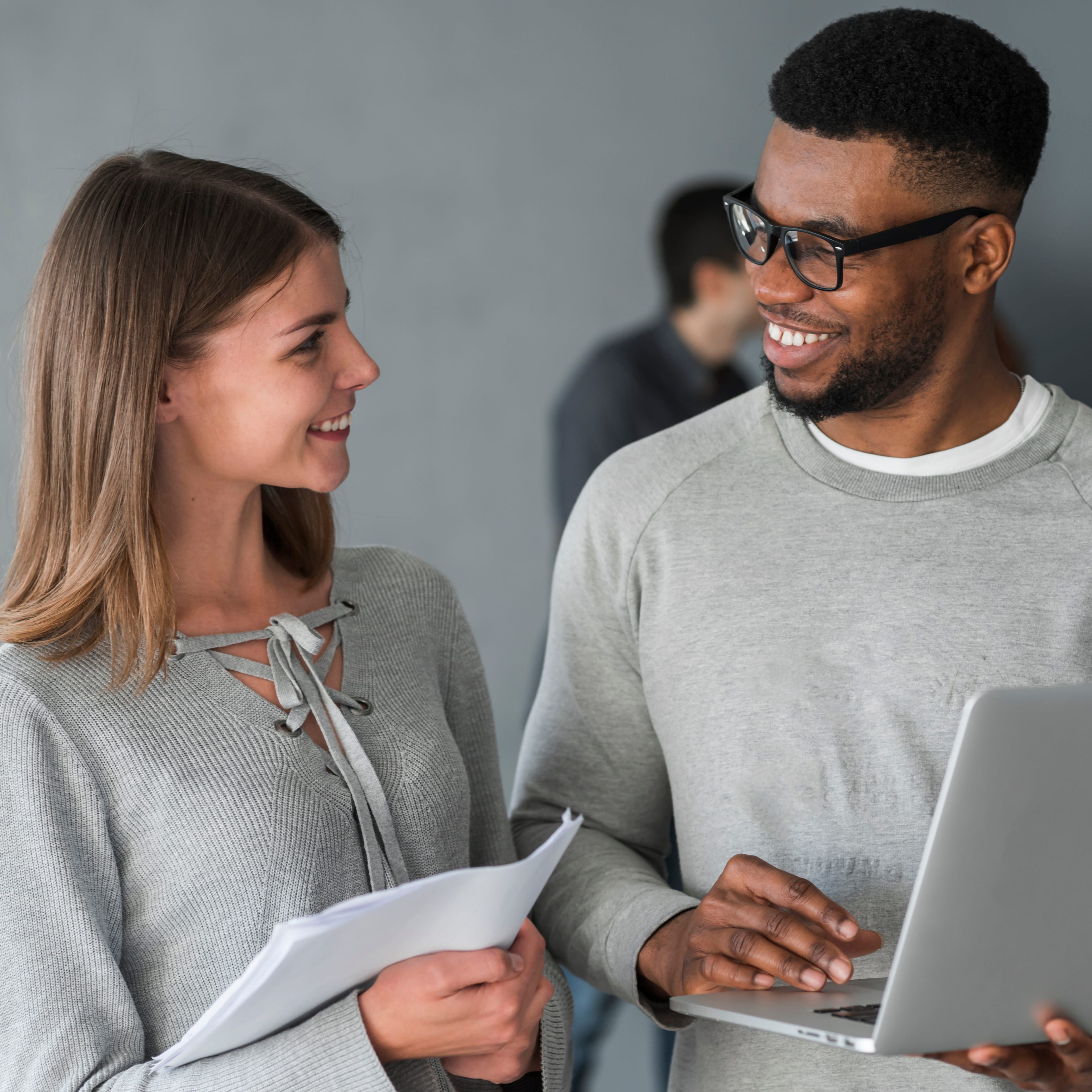 Two colleagues in conversation — one holding notes, the other showing something on a laptop.